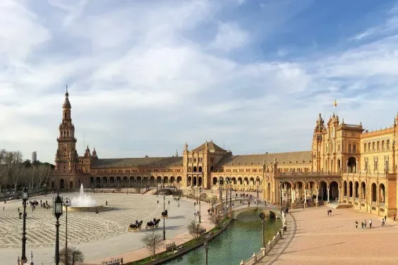 Travelers walking beside the canal and tiled alcoves of Plaza de España under a clear Seville sky